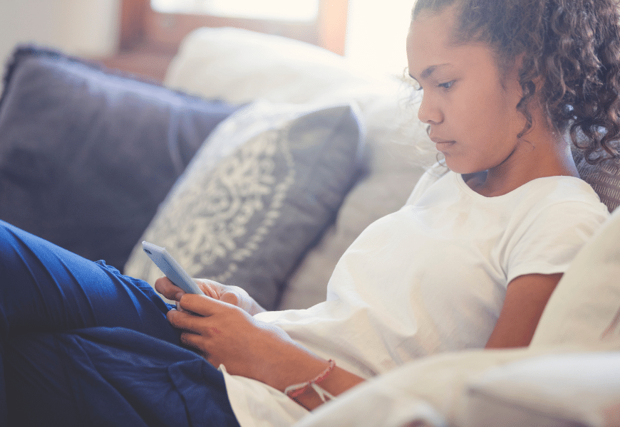 Teenager with long curly hair sitting on couch looking at mobile phone in her hands