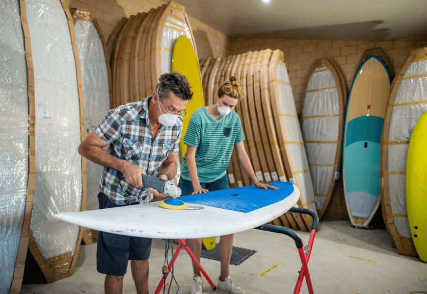 Two people in surfboard workshop sanding a surfboard
